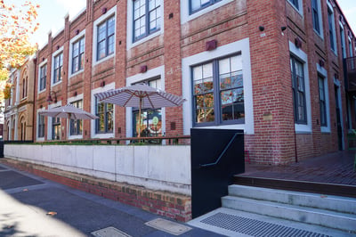 Red brick building, two levels high and large square windows with two market umbrellas out the front of the building.
